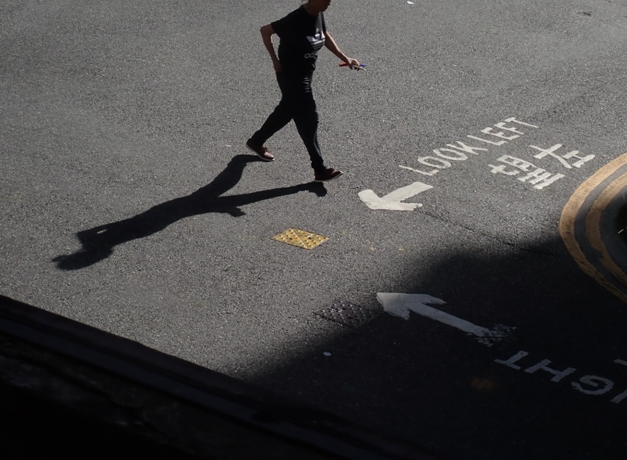 Une personne marche dans une rue de Hong Kong, projetant une longue ombre, avec des marquages routiers en chinois et en anglais.