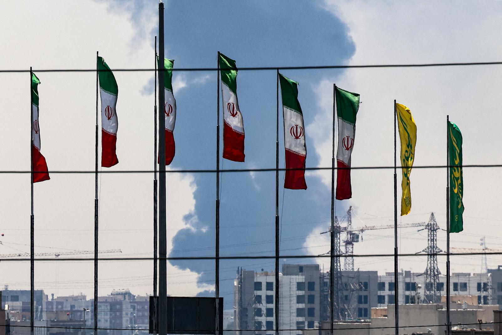 Des drapeaux iraniens à Téhéran, tandis qu'un panache de fumée s'élève d'une raffinerie de pétrole en arrière-plan, le 15 juin 2025.
