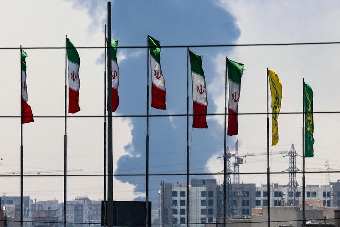 Des drapeaux iraniens à Téhéran, tandis qu'un panache de fumée s'élève d'une raffinerie de pétrole en arrière-plan, le 15 juin 2025.
