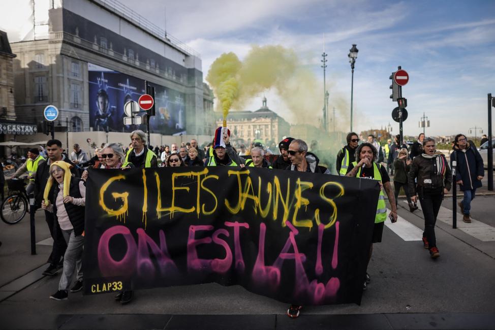 Des manifestants, vêtus de gilets jaunes, brandissent une fusée éclairante et une banderole sur laquelle on peut lire « Gilets jaunes, nous sommes là » lors d'un rassemblement marquant le 5e anniversaire des manifestations antigouvernementales dites « Gilets jaunes » de 2018-2019, à Bordeaux, en France, le 18 novembre 2023.