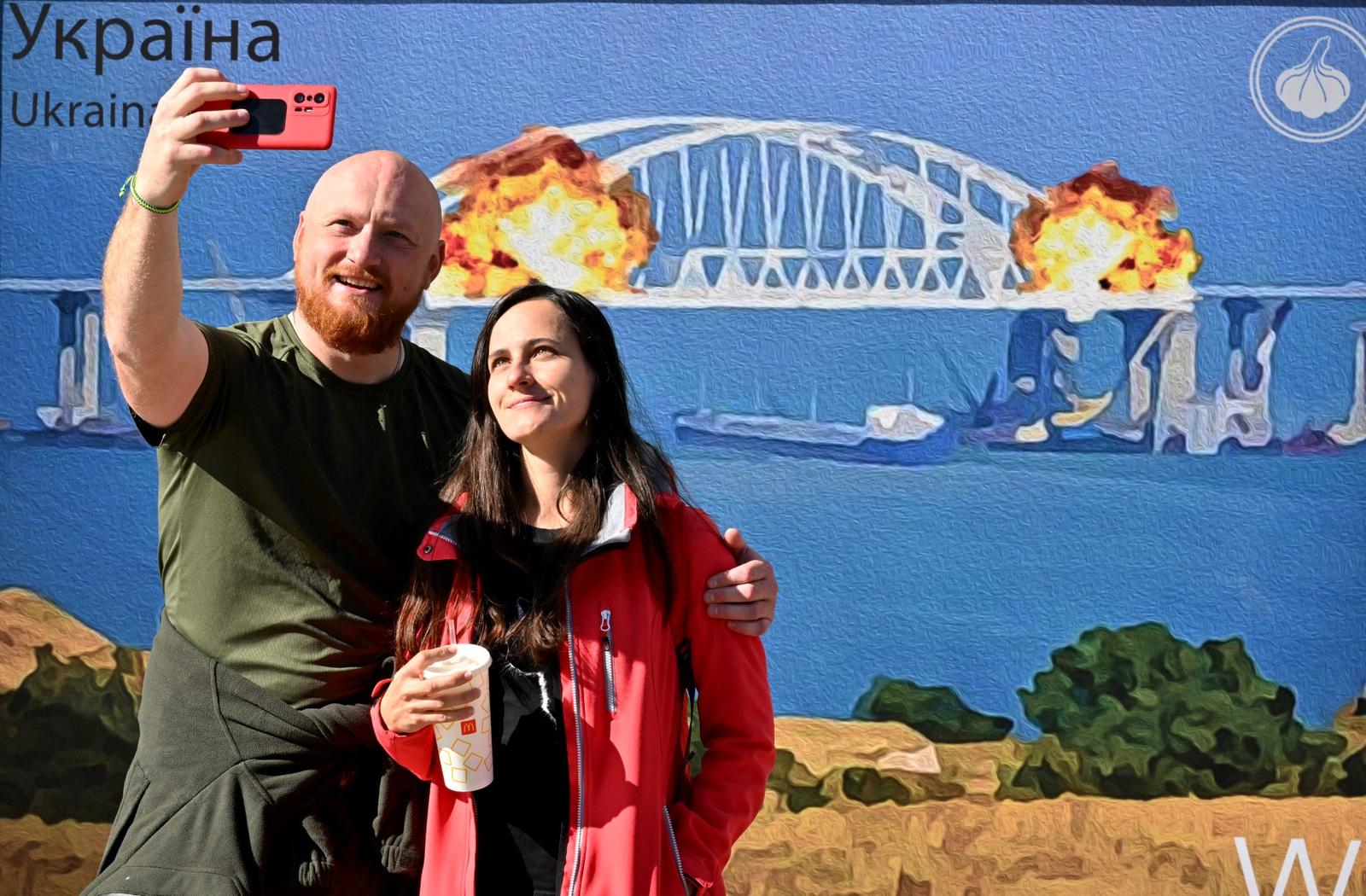 Un couple prend un selfie devant une œuvre d'art en forme de grand timbre intitulée « Du coton au pont de Crimée » créée par Andrusiv V., Serdyukov O., Kalinovska Y., Visich M. représentant une explosion sur le pont de Kertch