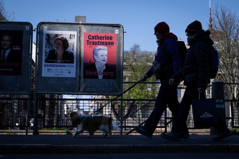 Des passants déambulent devant les affiches électorales de Jean-Philippe Vetter (à gauche), candidat des Républicains (LR) à la mairie de Strasbourg, de Jeanne Barseghian, maire sortante de Strasbourg et candidate à sa réélection, et de Catherine Trautmann (à droite), candidate du Parti socialiste (PS) à la mairie de Strasbourg, à la veille du second tour des élections municipales à Strasbourg, dans l'est de la France, le 18 mars 2026