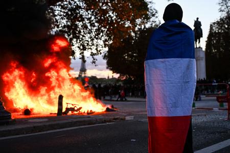 Un manifestant drapé dans le drapeau français se tient près d’un feu, avec la tour Eiffel en arrière-plan, lors d’une manifestation, à Paris, le 17 octobre 2026.